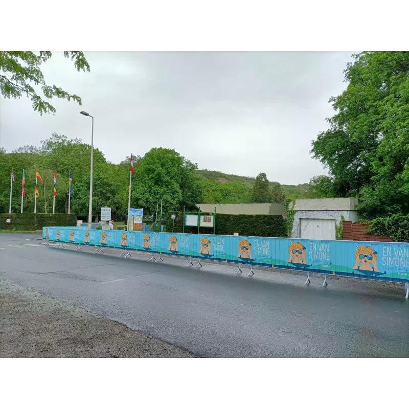 Barrières de sécurité avec banderoles publicitaires bleues affichant « EN VAN SIMONE » et mascotte, installées le long d’une route devant des arbres et un bâtiment.