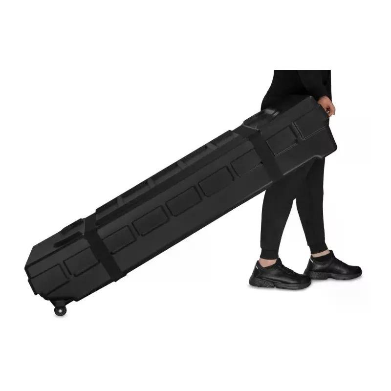 Homme transportant une grande valise noire rigide à roulettes, en position inclinée contre le corps.