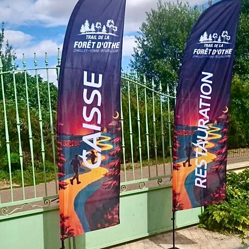 Tailles publicitaires en bannière tissu imprimé avec texte vertical « GUIDE » et logos Trail de la Forêt d’Ôthe, près d’une barrière, couleurs fortes et dégradé.